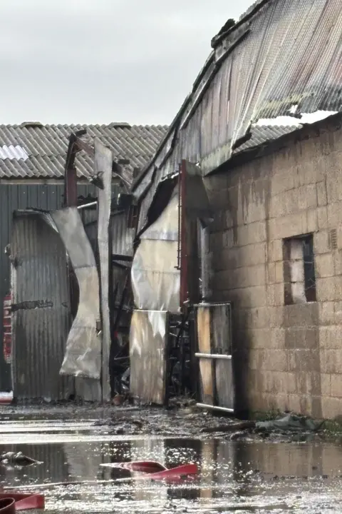 Megan Heaton A close-up of a fire-damaged building with a buckled roof, parts coming away and water around the walls after the firefighters put out the blaze.