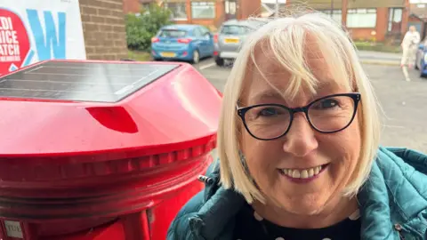 Woman with short blonde hair to her shoulders smiling. She is wearing black glasses and is wearing a poco-dot top and a blue coat. She is standing beside a red post-box which has a solar panel on its top. She is standing in a carpark.