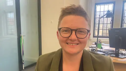 Bev Craig in an office, wearing an olive green blazer, glasses with tortoiseshell rims and short , light brown hair.