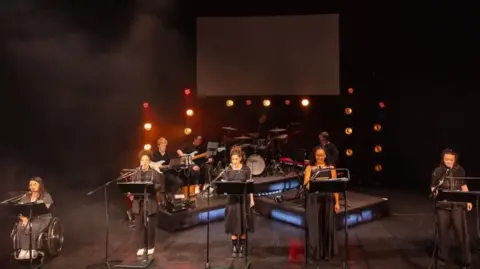Birmingham Hippodrome/Danny Kaan Five women lined up on a black stage singing. Behind them is a band and behind the band, there are stage lights. They are Elena Pitsiaeli, Courtney Stapleton, Elexi Walker, Maddison Bulleyment and Rosie Coles. 