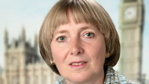 BBC/Flying Colours Councillor Deirdre Alden, she has medium length dark blonde hair. Behind her is a blurred image of the Palace of Westminster