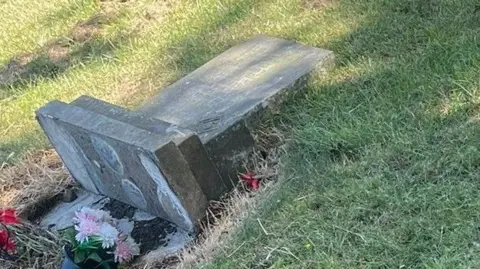Pat Grayson A knocked-over tomb stone, with flowers at the foot, in a grassed cemetery.