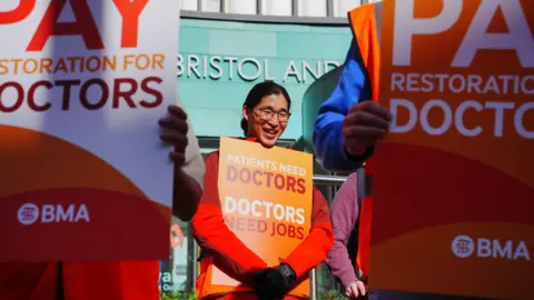 Ben Birchall/PA Wire An asian man is pictured holding up an orange sign which reads 'Patients need doctors. Doctors need jobs.' He is wearing glasses and smiling. Other people are around him also holding the same sign but he is in focus. He is standing in front of the Bristol Royal Infirmary on a sunny day.