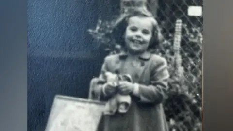 Maggie Hackney A black and white photo of Maggie Hackney as a young child. She stands outside in front of a wire fence, holding a pair of sandals and a shopping back. She has chin length hair and is smiling widely. She wears a smart duffel coat with a collar and double breasting.