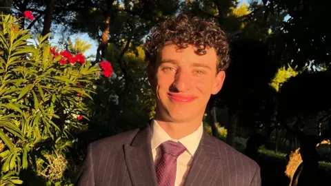 Daniel Daniel, who has dark curly hair and is wearing a purple suit stood outside with green bushes and red flowers in the background. 