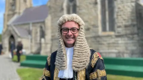 Juan Watterson, a man who wears glasses smiles brightly, he wears a long white wig and decadent black and gold robes as he stands in front of an old Church at Tynwald.