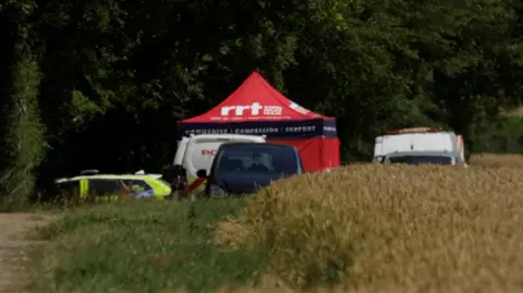 Jamie Niblock/BBC Vehicles belonging to the police and other emergency workers parked by the side of the field, where a red tent has been set up.