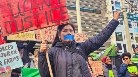 United Voices of the World Julia Quecano Casimiro holding a red protest sign , saying "we want justice", while punching the air and surrounded by fellow former farm workers