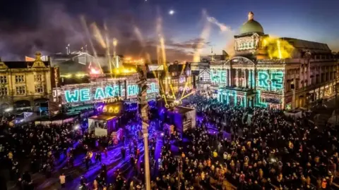 An event in Hull city centre in 2017. The words "we are Hull" are projected onto key city centre buildings. There are gold and purple lights which shine on a large crowd gathered in Victoria Square. 