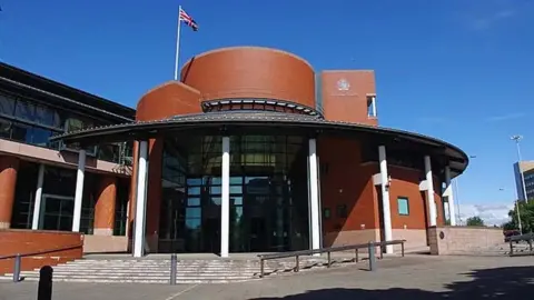 Ian Taylor/Geograph External view of the entrance to Preston Crown Court, a round red brick building shaped like a hat with columns supporting the round roof and steps up to the front against a brilliant blue sky