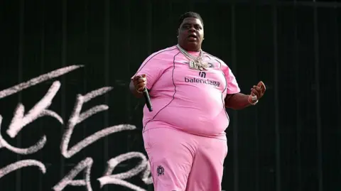 The rapper wearing a pink outfit with 'balenciaga' written on the chest, and several necklaces, holds a microphone in his extended right hand away against a black background with some chalk-like letters to his right 
