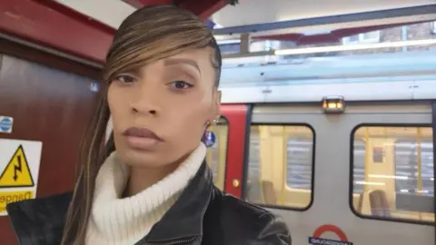 Cynthia Nodanche Cynthia Nodanche in London. She is standing in front of a tube train. She has a long fringe and is taking a selfie. 