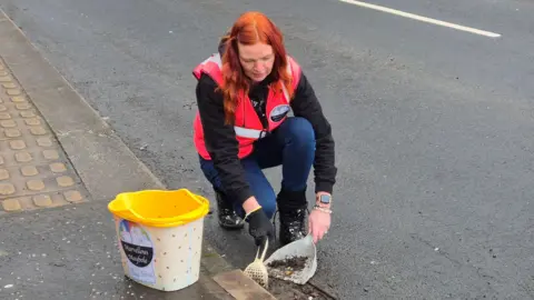 A woman wearing a pink high vis vest and jeans, shovelling up dirt by the side of a road. A large bucket with yellow trim is next to her.