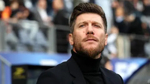 A man wearing a black polo neck and blazer with a quiffed hairstyle and a beard. He is standing in front of a stand full of football fans and is gazing into the distance.