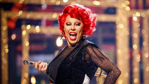 BBC/Guy Levy La Voix in drag at Strictly with bright red hair and lipstick and a black sparkly gown.