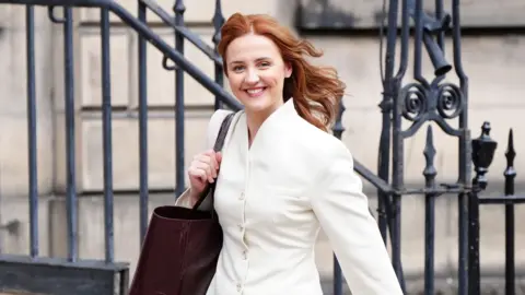 PA Media Culture Secretary Màiri McAllan, a young woman with red hair, smiles for the camera as she walks into Bute House, the Edinburgh Residence of the first minister. She wears a white blazer and carries a dark brown leather tote bag
