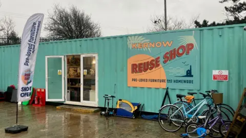 Bright green shipping container full of stuff for sale at Dump in Falmouth. A sign on the building reads 'Kernow Reuse Shop - pre-loved furniture and homewear. Two bikes and three scooters plus a bike trailer are stood up outside the container to the right of a white uPVC patio door leading inside.