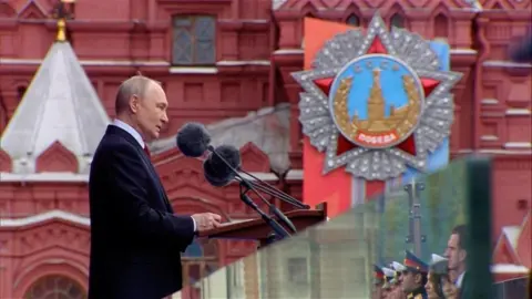 Russian President Vladimir Putin stands wearing a dark suit at a lectern in Moscow's Red Square as he gives a speech.