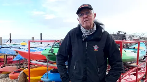 Ian Cobb is on the right of the frame, wearing a cap and a pair of glasses. He is wearing a scarf, a Southampton Football Club rain jacket and blue jeans. Behind him are rows of kayaks and canoes in different colours, and in the background is the Bristol channel.