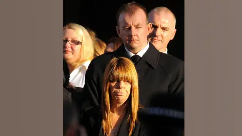 Hazel Barker pictured wearing black at the funeral of her husband PC Bill Barker in 2009. She has long, straight red hair with a straight fringe. Two men and a woman can be seen clearly behind her, among a larger group of mourners.