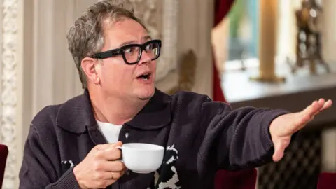 Studio Lambert/BBC/PA Alan Carr in the breakfast room on The Celebrity Traitors. He has greying hair and is wearing thick black framed glasses and a dark purple collared jumper. In his right hand is a white cup and his left is out stretched.