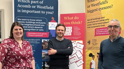 A woman and two men stand in front three colourful display boards, about Acomb and Westfield suburbs. From left to right, the woman has short brown hair in a bob and wears a red blouse with a pattern of white leaves on it. The man has short brown hair, a brown beard and wears a navy blue jumper. The man on the right has grey hair and glasses, and wears a navy blue jumper.