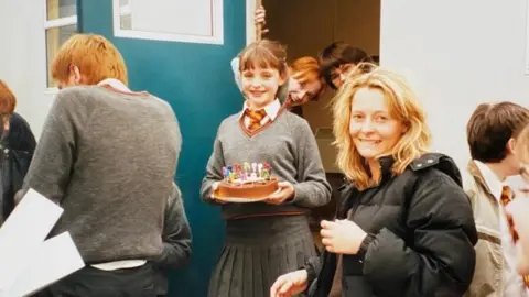 A young girl dressed in a Hogwarts school uniform is smiling holding a birthday cake. She is surrounded by people, some resembling characters from the Harry Potter movies.