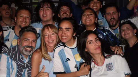 A group of football fans gathered to watch a World Cup match wearing mainly Argentinian football shirts. The woman on the front row far right is wearing an England shirt. They are in a pub and are all smiling.