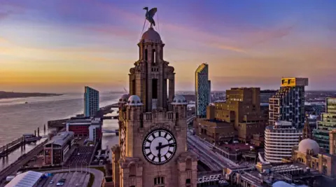 A view of the clock and Liverbird on the Liverpool Liver building with the River Mersey in the background taken at sunrise. A number of high-rise buildings are in the background.