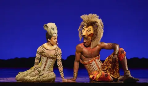 Matt Crockett A female and male actor sit on stage dressed in costumes from the Lion King. They are turned toward one another, as if mid conversation.
The woman is wearing a lioness mask perched on the top of his head, tight fitting costume in shades of brown and beige, and her arms are painted in brown and white cross patterns. The man on the right wears a lion mask with make atop his head, a red, yellow, brown and white corset with skirt, and has ankle and arm bracelets that appear bronze in colour. Behind them is a dark blue backdrop representing night on the Savannah.