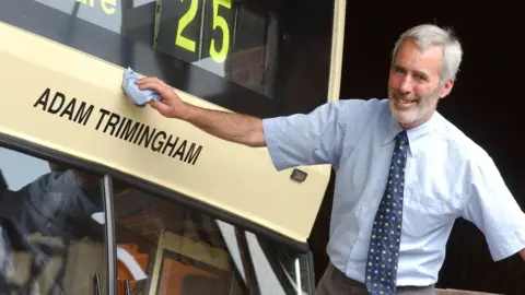 Adam Trimingham - a man in a blue shirt and tie polishes a yellow bus with his name on the front