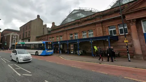 An exterior view of Carlisle Market Hall, with a bus shelter on the external wall. a Bus waits in the bus stop and people are walking on the pavement nearby.