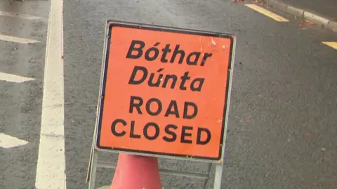 An orange road closed sign written in English and 'Bóther Dúnta' in Irish is placed in the middle of the road.