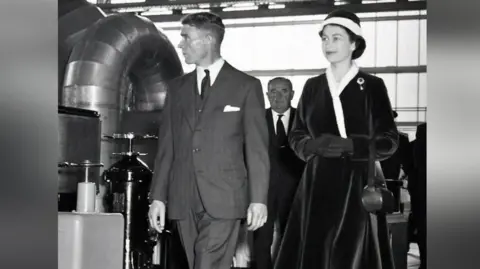 NDA A black and white image of a young Queen Elizabeth II walking inside Calder Hall next to a man in a suit with a white handkerchief peeping out of his top pocket. The Queen is wearing a dark velvet dress and matching hat. A larger, bald man is behind her and her companion, seen through the gap between them.