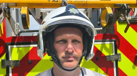 A firefighter wearing his uniform, looking straight at the camera standing at the back of a fire engine.