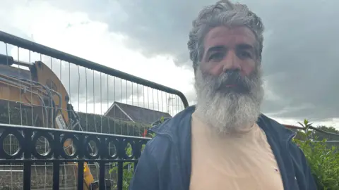 Julia Gregory / BBC A man standing next to a metal railing fence with a digger working behind him. He has grey hair and a grey beard, and wearing a beige top and a blue jacket