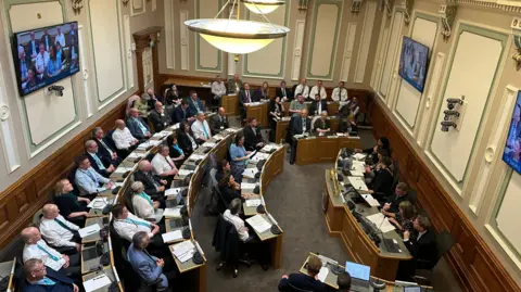 Looking down into a council chambers, circular rows of desks with people sat in smart clothes. In front of them a smaller desk with five people sat facing them. Screens are on the walls of the room and there are overhanging lights.