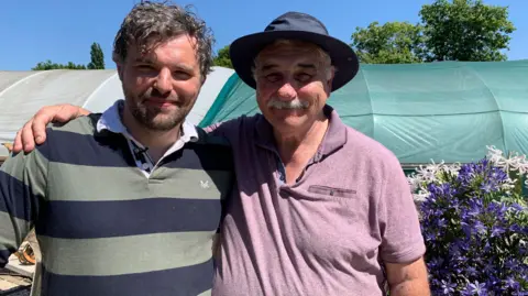 Two men with their arms around each other standing outside in the sunshine. The man on the left is younger and is wearing a long-sleeved striped top while the man on the right is wearing a dark pink T-shirt and a hat.