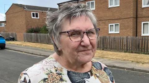 Kate Bradbrook/BBC Jan Donald, de pie en el exterior, con el pelo corto y gris y gafas. Lleva una blusa estampada de flores. Está junto a una casa con una valla de madera delante.