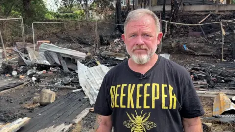 BBC/NATHAN TURVEY Richard Terry stands in the charred remains of the allotments. He has grey hair and a grey beard and is wearing a black T-shirt with the text "beekeeper" on it and a picture of a bee. There is twisted mental and burnt wood all over the floor