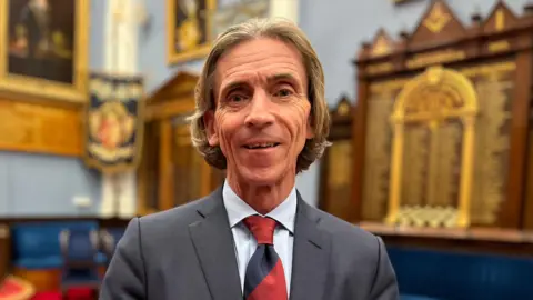 Tim Dallas-Chapman, head of the Jersey Freemasons, is dressed in a grey suit with a blue shirt and a navy and red tie. He is standing in an elegant looking room with lots of old plaques on the wall. He is looking directly at the camera, smiling.  