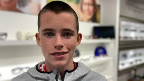 BBC A boy with short brown hair and blue eyes is smiling at the camera. He is wearing braces on his teeth and has a grey hoodie top on.