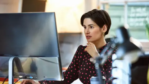 Getty Images A woman sits at a desk looking at a computer monitor with a slightly worried look on her face.