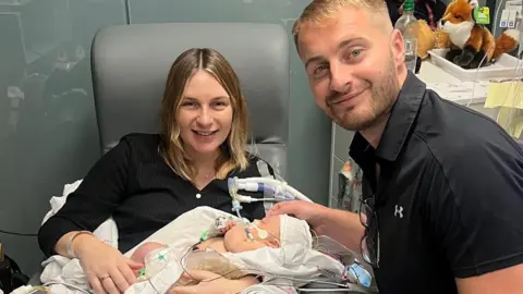Victoria and Thomas Gillibrand with baby Pippa. Victoria has blonde hair and is wearing a black top. She is cradling the baby which has multiple tubes attached to her. Thomas Giillibanrd has a black short sleeved shirt and has his hand on the baby's head.