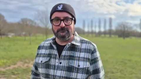 Stuart Woodward/BBC Aaron Davies wears a black and white checked shirt, open at the neck, with a black T-shirt underneath. He is wearing a black beanie cap with a white dog design on the front. Aaron has dark-rimmed glasses and a dark beard. He is standing on an area of grass with trees in the background and blue sky above