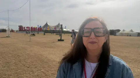 A woman with long dark hair and thick-rimmed glasses wearing a white t shirt and denim overshirt. She is stood in a large field with tents and a large Eisteddfod sign in the background. She is looking at the camera and smiling.
