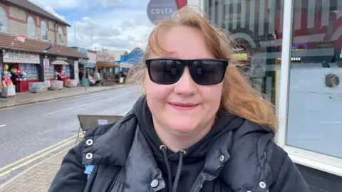 A head and shoulders shot of a woman with brown hair tied back, black sunglasses, a black hoodie and coat. Behind her are seaside shops with colourful beach balls out the front and a Costa sign.