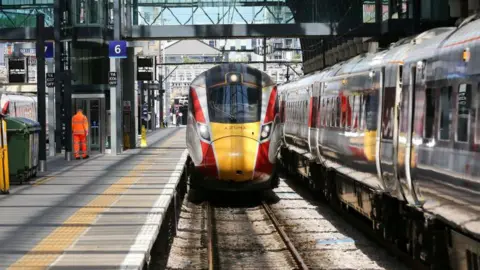 A modern high-speed train with a yellow and red front is stopped at a railway platform under a glass and metal station roof.

Another long train runs parallel on the right, reflecting the surroundings on its shiny exterior.

Platform signs, safety markings, and a worker in bright orange clothing are visible along the station.