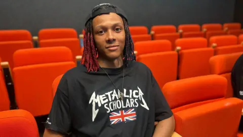 Kysharn Queensborough looks to camera. He wears a Metallica Scholars T Shirt. 