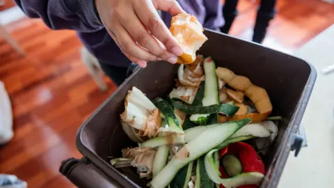 Close up of a person dropping an apple core into a small kitchen caddy bin full of vegetable peelings